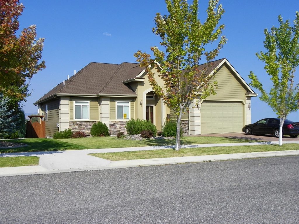 Tan two story home with a double garage and young trees along a quiet residential street